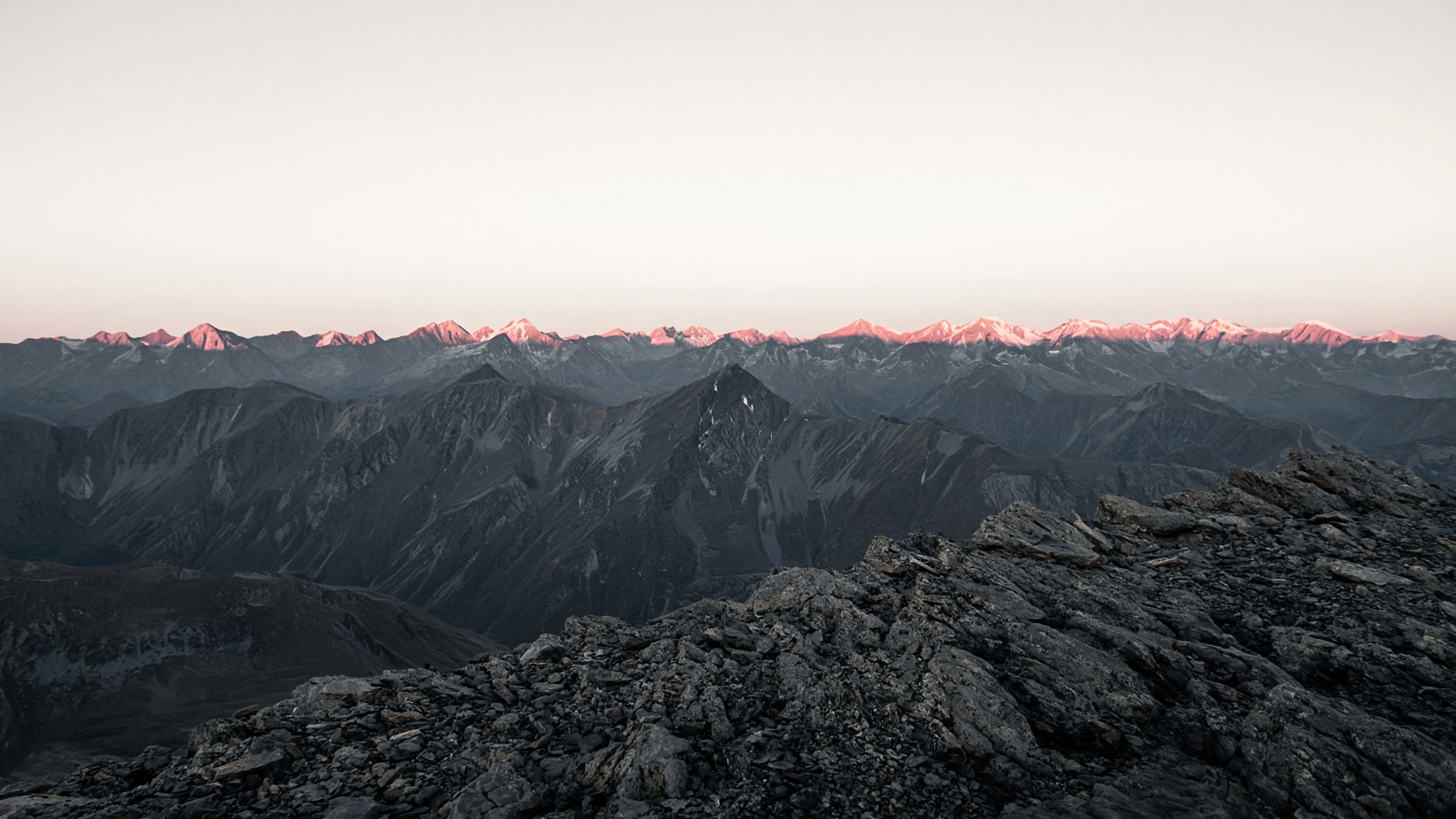 A high ridge at first light, stone in the foreground and a thin rose line on the far peaks.